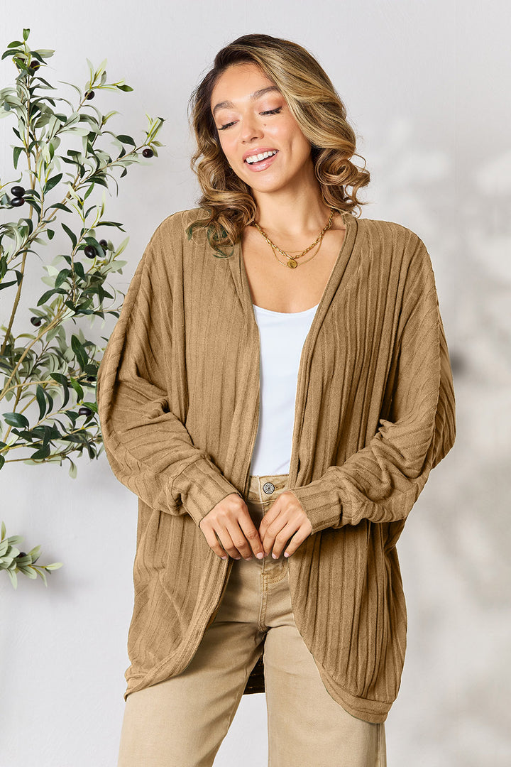 A person with long, wavy hair is smiling and looking away from the camera while embracing a basic style. They are wearing the Basic Bae Full Size Ribbed Cocoon Cardigan in beige over a white top made of slightly stretchy polyester, paired with light-colored pants. A leafy plant is partially visible in the background.