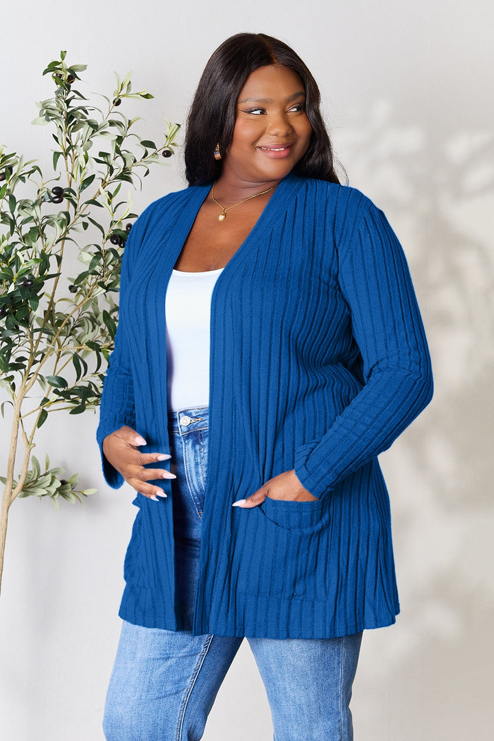 A curve model stands smiling, wearing the Basic Bae Full Size Ribbed Open Front Cardigan with Pockets in blue over a white top, paired with comfy blue jeans crafted from a pocketed stretch polyester elastane blend. Next to them, a lush green indoor plant adds vibrancy against the light-colored wall.