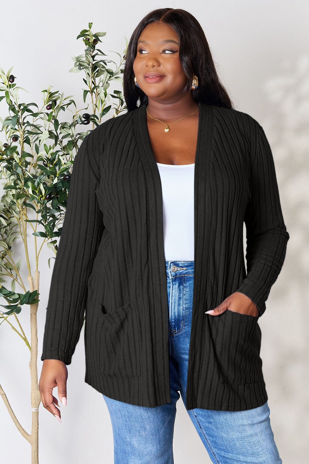 A curve model stands confidently, donning the Basic Bae Full Size Ribbed Open Front Cardigan with Pockets in black, made from a polyester elastane blend, over a white top and blue jeans. Her hands rest casually in the pocketed stretch of her cardigan. In the background, a vibrant plant with green leaves adds a touch of nature to the scene.