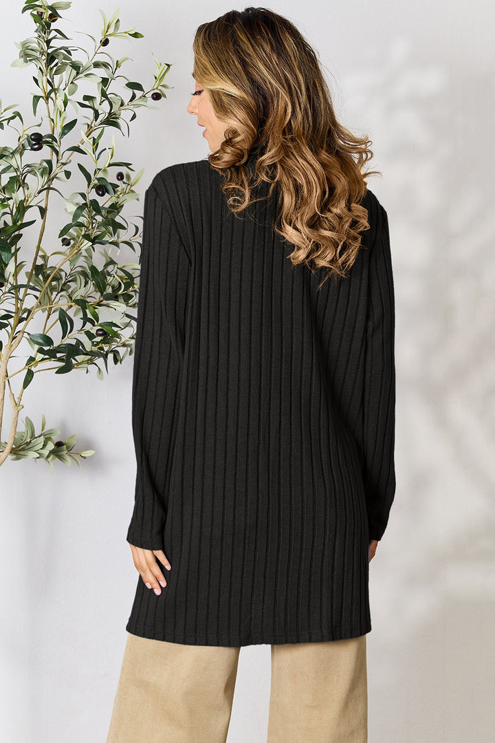 A person with long wavy hair wears a black Basic Bae Full Size Ribbed Open Front Cardigan with Pockets and beige, pocketed stretch pants, standing with their back to the camera. On the left, an olive plant with black olives is visible in the background.