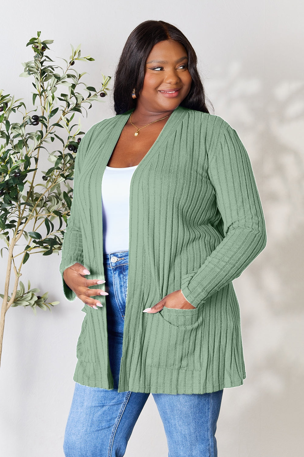 A woman with long, dark hair smiles beside a leafy plant, wearing jeans and the "Basic Bae Full Size Ribbed Open Front Cardigan with Pockets" in green. She has layered it over a white top, enjoying both comfort and style. The background is a plain, light color.