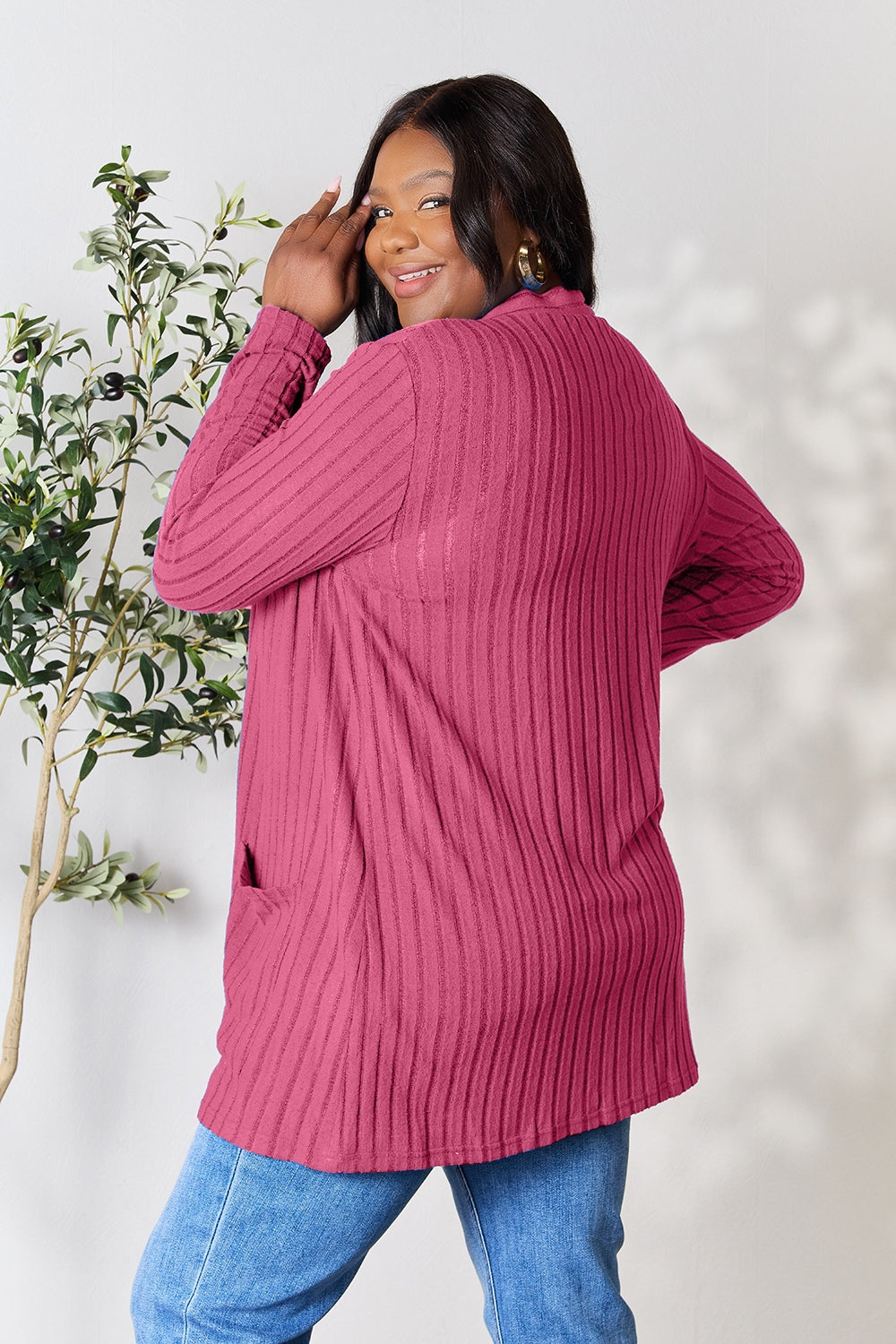 Dressed in the Basic Bae Full Size Ribbed Open Front Cardigan with Pockets, a person wearing pink and blue jeans poses playfully against a light background with a nearby potted plant. The photo captures their plus-size outfit's comfortable pocketed stretch as they smile and look backward.