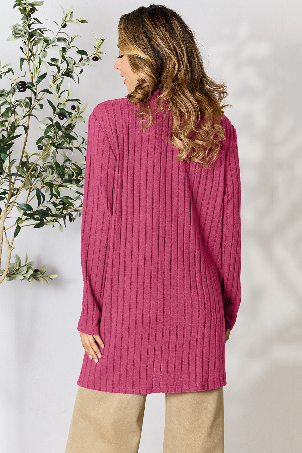 A woman with long, wavy hair stands with her back to the camera, wearing the cozy Basic Bae Full Size Ribbed Open Front Cardigan with Pockets in magenta over beige pants. The plus-size design drapes gracefully beside a leafy plant against a neutral background and is machine washable.