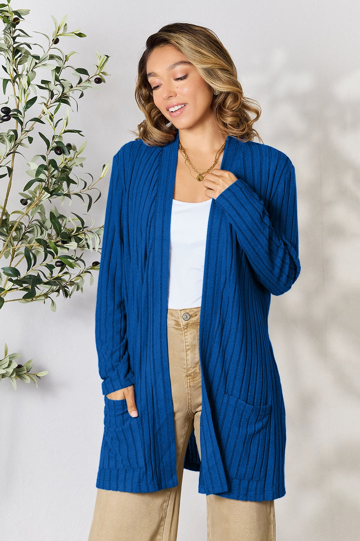 A person stands near an olive plant, dressed in the Basic Bae Full Size Ribbed Open Front Cardigan with Pockets in blue, layered over a white top and paired with beige pocketed stretch pants. Their wavy hair cascades as they look down with a gentle smile. The scene is set against a plain wall illuminated by natural lighting.