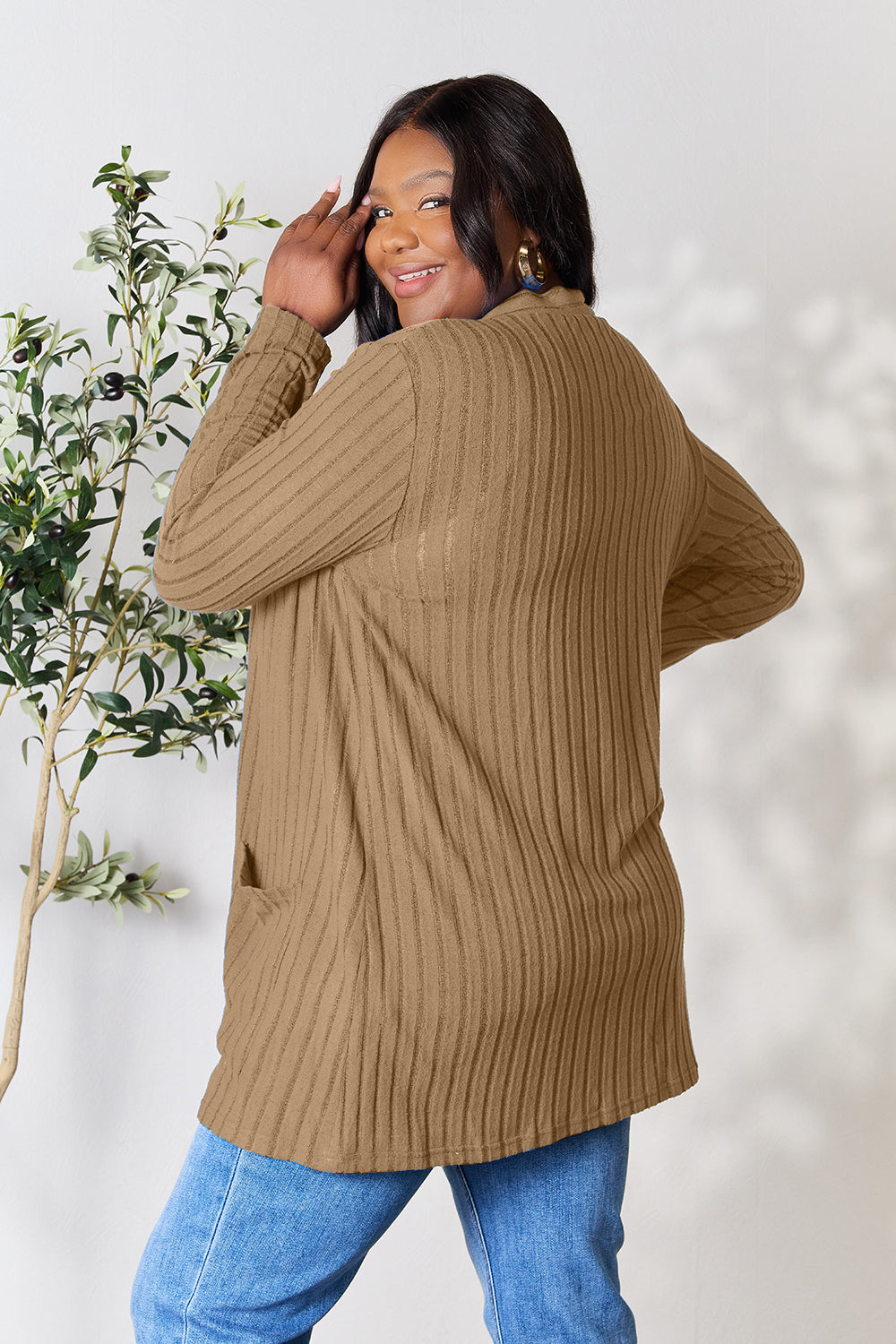 A woman with long dark hair smiles and glances over her shoulder, wearing the Basic Bae Full Size Ribbed Open Front Cardigan with Pockets in a camel color, paired with blue jeans. Her machine-washable outfit emphasizes convenience and easy care. In the background, a plant with green leaves adds a touch of nature to the scene.