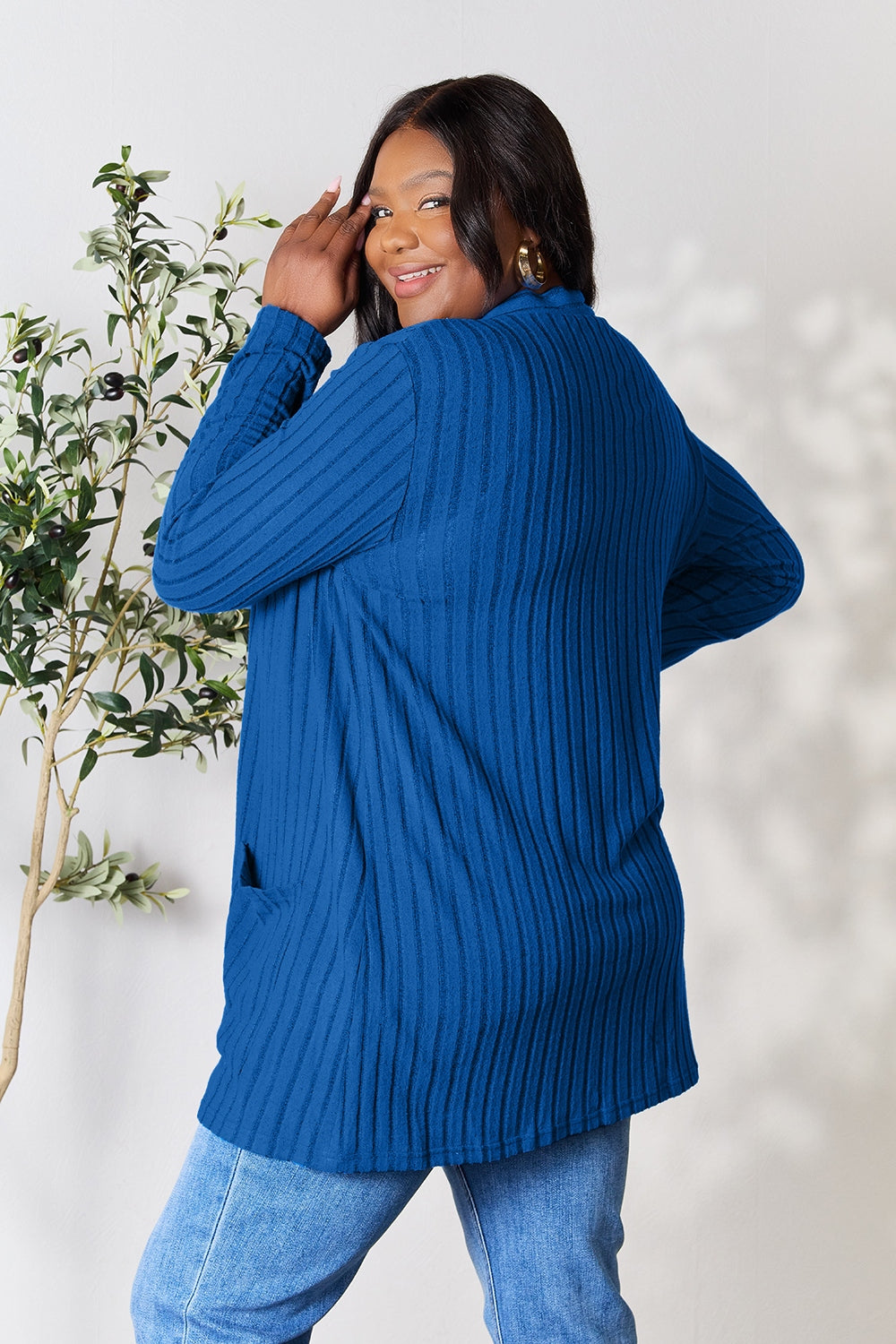 A woman in the Basic Bae Full Size Ribbed Open Front Cardigan with Pockets, paired with jeans, smiles over her shoulder while standing near a small tree against a light background.