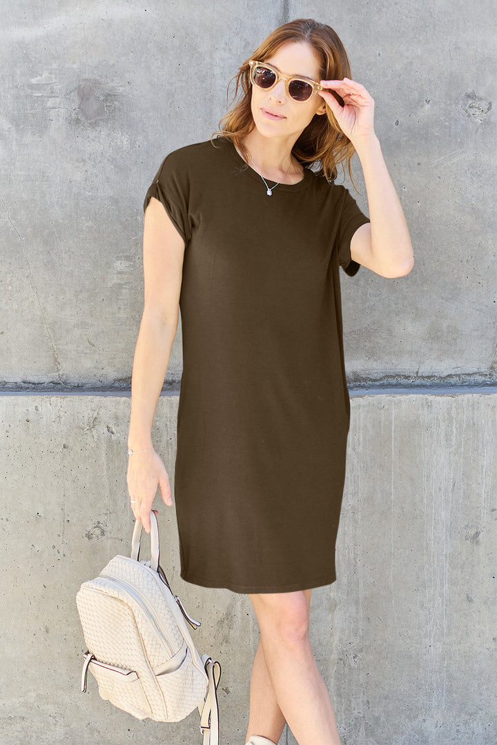 A person with long hair in sunglasses and a casual black Basic Bae Full Size Round Neck Short Sleeve Dress with Pockets leans against a concrete wall. They hold a textured polyester backpack in one hand and adjust their sunglasses with the other.