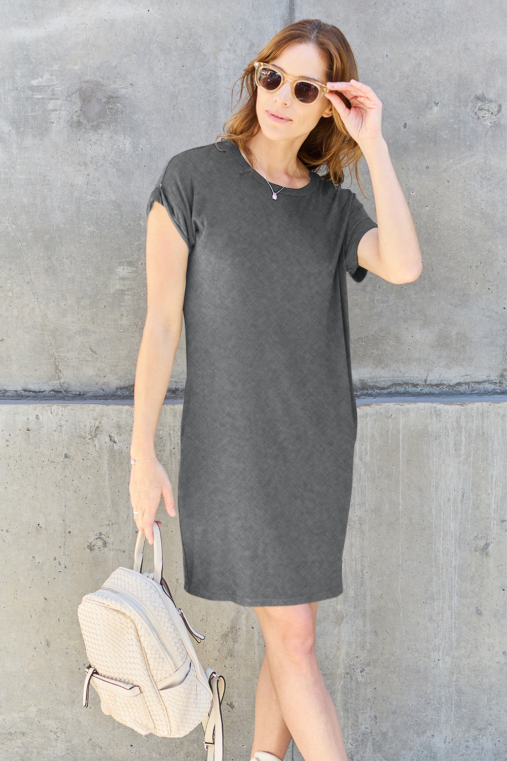 A woman in a Basic Bae Full Size Round Neck Short Sleeve Dress with Pockets in gray, along with sunglasses, stands against a concrete wall. She's holding a cream-colored backpack and adjusting her sunglasses with her right hand.