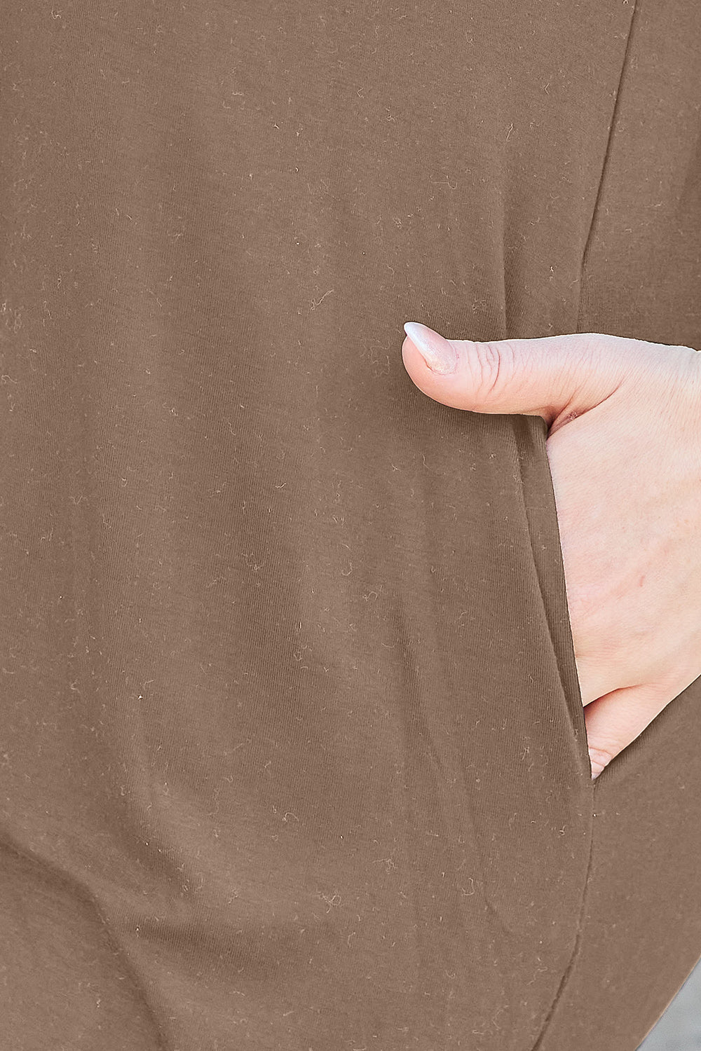 A close-up showcases a hand partially tucked into the pocket of a Basic Bae Full Size Round Neck Short Sleeve Dress. This brown garment seems to be crafted from a textured, soft blend, possibly polyester. The image highlights the dress's color and texture, indicating it might need delicate care such as machine washing in cold water.