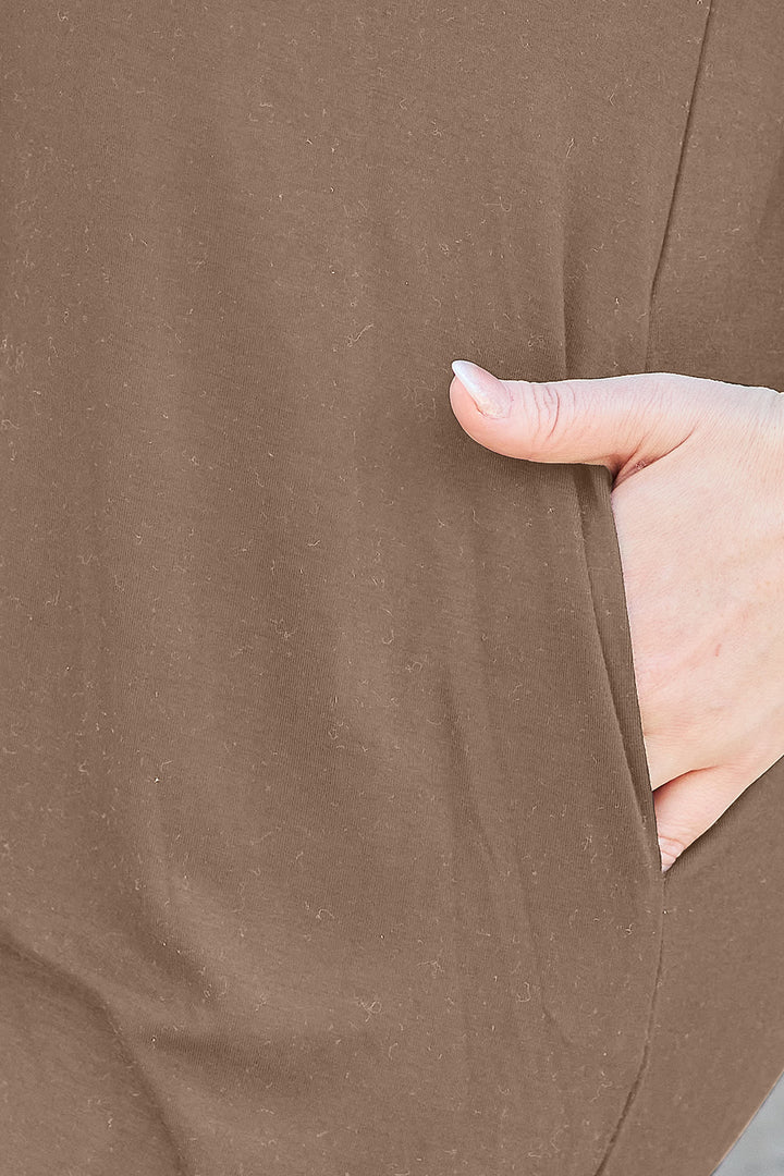 A close-up showcases a hand partially tucked into the pocket of a Basic Bae Full Size Round Neck Short Sleeve Dress. This brown garment seems to be crafted from a textured, soft blend, possibly polyester. The image highlights the dress's color and texture, indicating it might need delicate care such as machine washing in cold water.