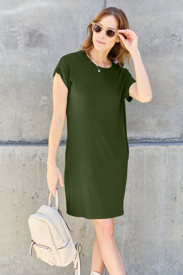 A person wearing sunglasses and the Basic Bae Full Size Round Neck Short Sleeve Dress with Pockets, crafted from a soft polyester and spandex blend in green, poses against a concrete wall while holding a light-colored backpack.