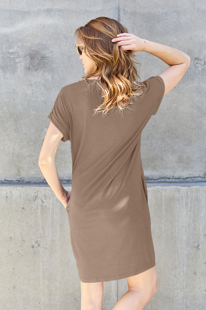 A person with long hair stands against a concrete wall, modeling the Basic Bae Full Size Round Neck Short Sleeve Dress with Pockets, crafted from soft polyester in a brown hue. Sunglasses conceal their eyes as they turn away from the camera, one hand resting on their head. This stylish ensemble provides everyday elegance and easy care with a simple machine wash cold.