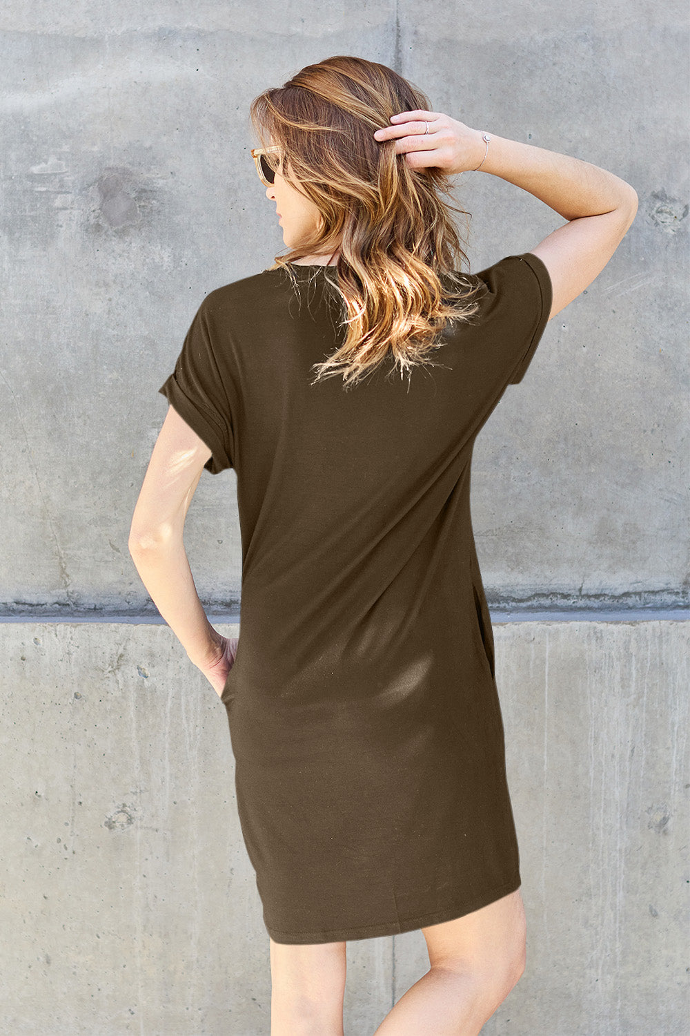 A woman with light brown hair, her back facing the camera, is wearing sunglasses and a Basic Bae Full Size Round Neck Short Sleeve Dress with Pockets in dark olive green. She stands against a concrete wall, one hand in her pocket while the other adjusts her sunglasses.