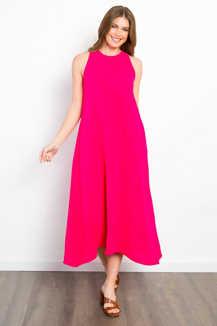 A person in a bright pink Be Stage Midi Tank Dress with Pockets stands on a wooden floor against a white background. Smiling with long hair and one arm slightly raised, their versatile outfit is complemented by casual brown sandals.