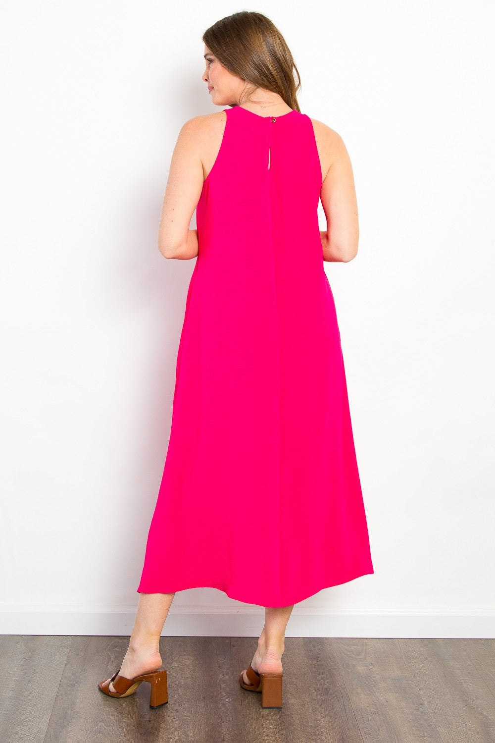 A woman in a vibrant pink Be Stage Midi Tank Dress with Pockets stands on a wooden floor, facing away. With light brown hair and brown heeled sandals, she showcases a versatile wardrobe choice against the plain white wall.