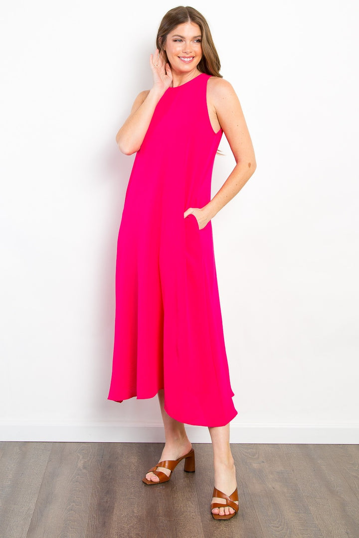 A person in a bright pink Be Stage Midi Tank Dress with pockets stands on a wooden floor against a white background. They smile, one hand touching their face, the other in their dress pocket, paired with brown sandals and long hair cascading.