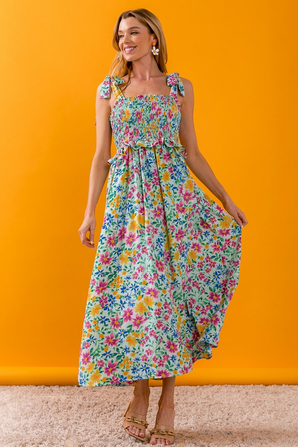 A woman wearing the BiBi Floral Ruffle Trim Smocked Cami Dress stands against an orange background, smiling. Her long, sleeveless dress with bow straps features vibrant shades of blue, pink, yellow, and green. She is wearing sandals and holds one side of her ruffle-trimmed dress out slightly.