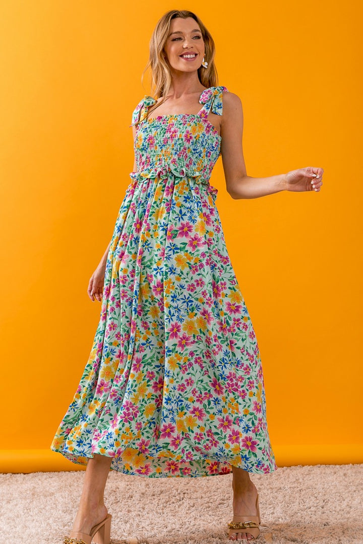 Modeling in front of a vivid orange backdrop, a person strikes a joyful pose in the BiBi Floral Ruffle Trim Smocked Cami Dress, showcasing its colorful floral design, elegant ruffle trim, and tied shoulder straps. The long, flowy dress beautifully complements their beaming smile.