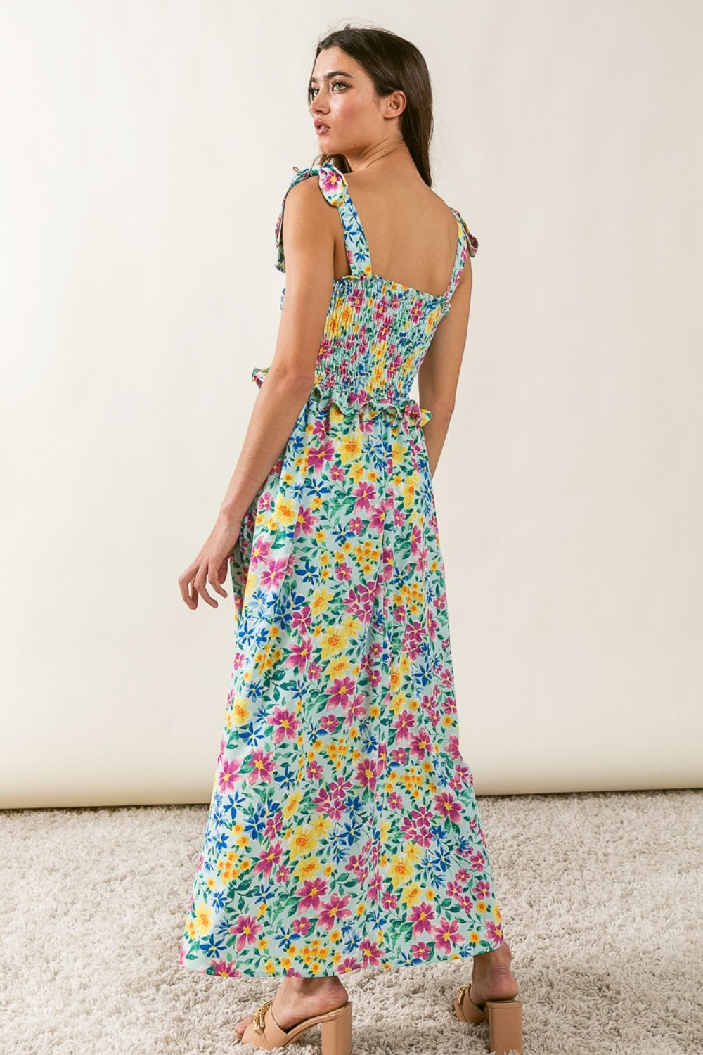 A woman stands on a carpeted floor, slightly turned away from the camera. She is dressed in the BiBi Floral Ruffle Trim Smocked Cami Dress, featuring tie straps, and pairs it with light-colored sandals. The backdrop is a plain, light-colored wall.