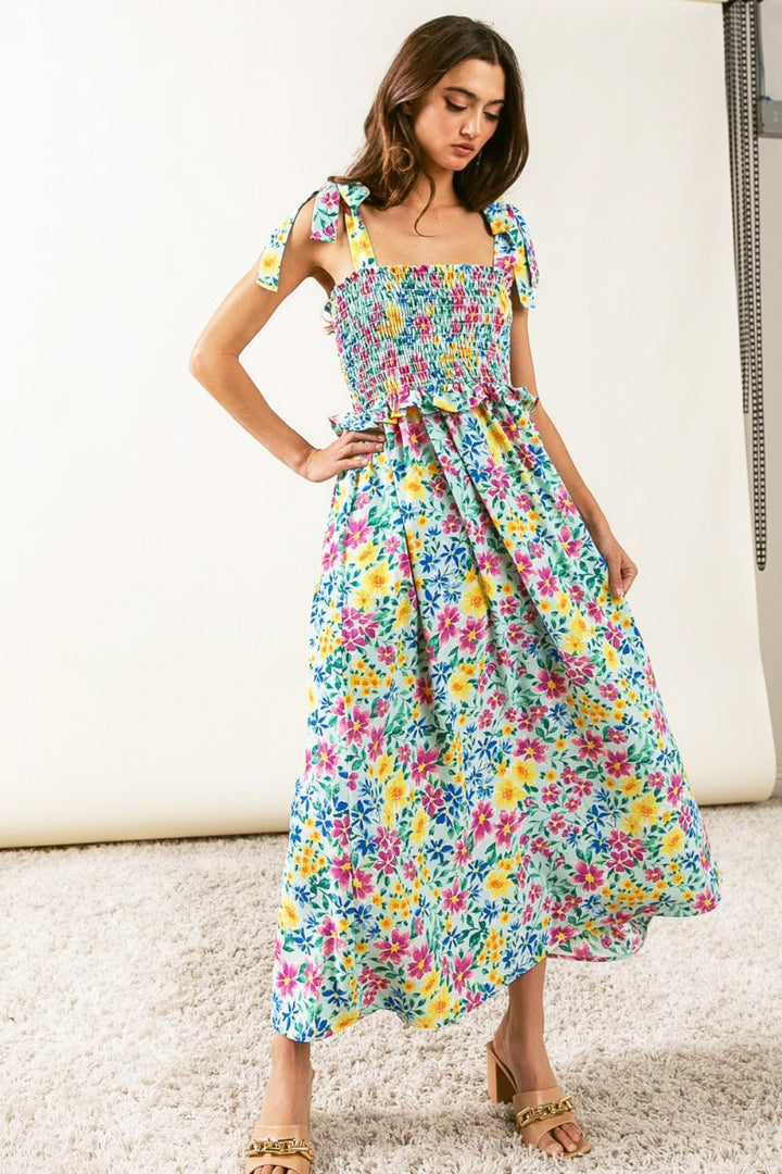 A woman stands on a carpeted floor, dressed in the BiBi Floral Ruffle Trim Smocked Cami Dress, featuring tied shoulder straps and a charming ruffle trim. This ankle-length dress pairs beautifully with her beige sandals against the backdrop of a plain, light-colored wall.