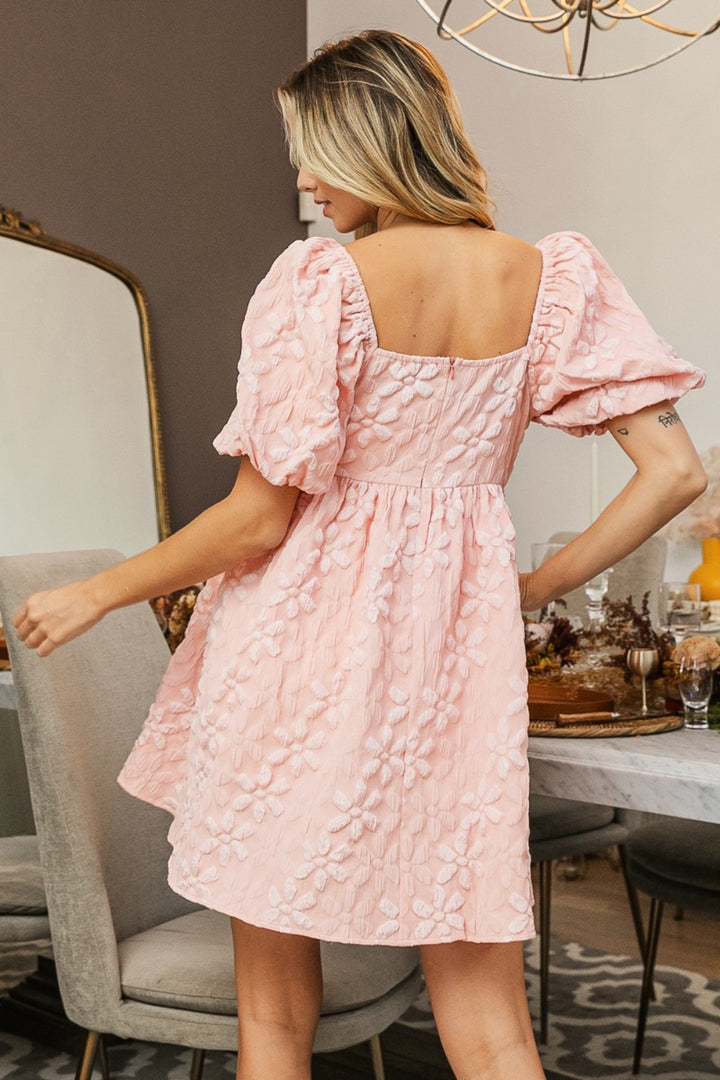 A woman stands in the BiBi Flower Square Neck Puff Sleeve Dress, showcasing its romantic design with pink, textured puff sleeves and a square neckline. Her long blonde hair cascades as she poses in a room decorated with a dining table adorned with glassware and candles, beside a large mirror on the wall.