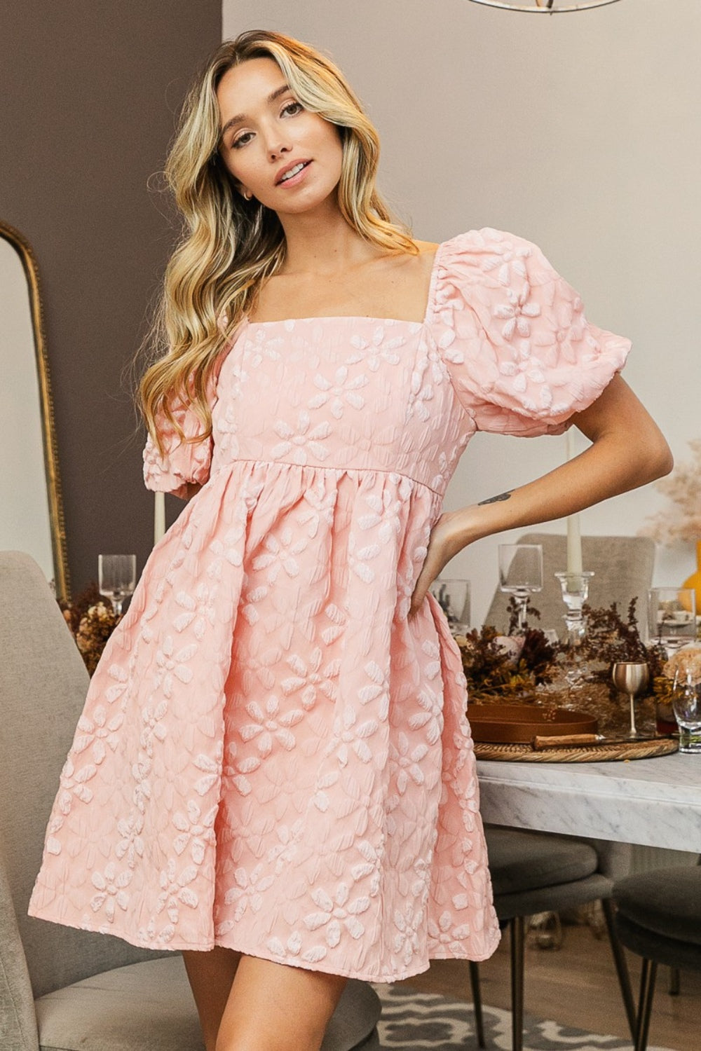 A woman wearing the BiBi Flower Square Neck Puff Sleeve Dress in light pink, adorned with floral details, stands in a dining room. The elegantly furnished room features a table set for a meal, complete with glassware and decorative elements. Her long, wavy blonde hair perfectly complements her enchanting attire.