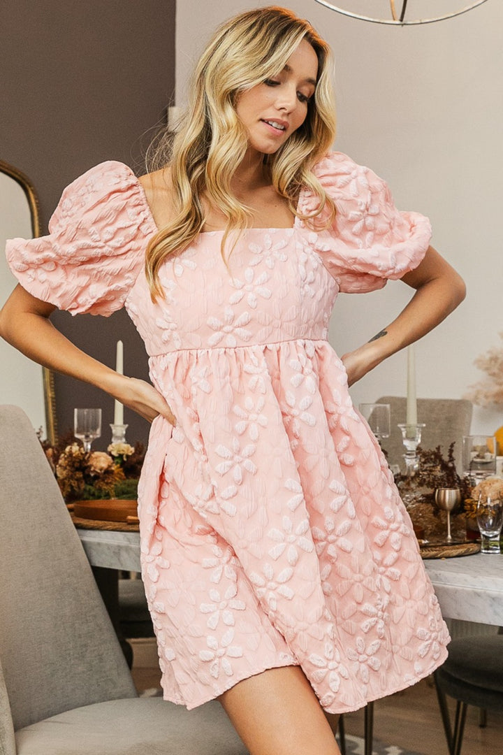 A woman clad in the BiBi Flower Square Neck Puff Sleeve Dress, a romantic pink attire, stands indoors. With long, wavy blonde hair cascading down her shoulders, she is positioned near an elegantly set dining table adorned with candles and decorative arrangements. The setting exudes sophistication and warmth, further enhanced by floral details that add to the overall inviting ambiance.