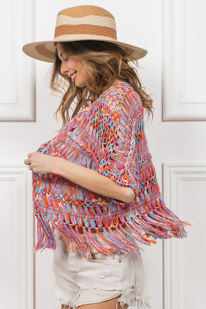 A woman in a straw hat models the BiBi Open Front Fringed Crop Knit Cardigan. Her wavy hair cascades over light denim shorts, highlighting the cardigans colorful crochet design against simple white paneled walls.