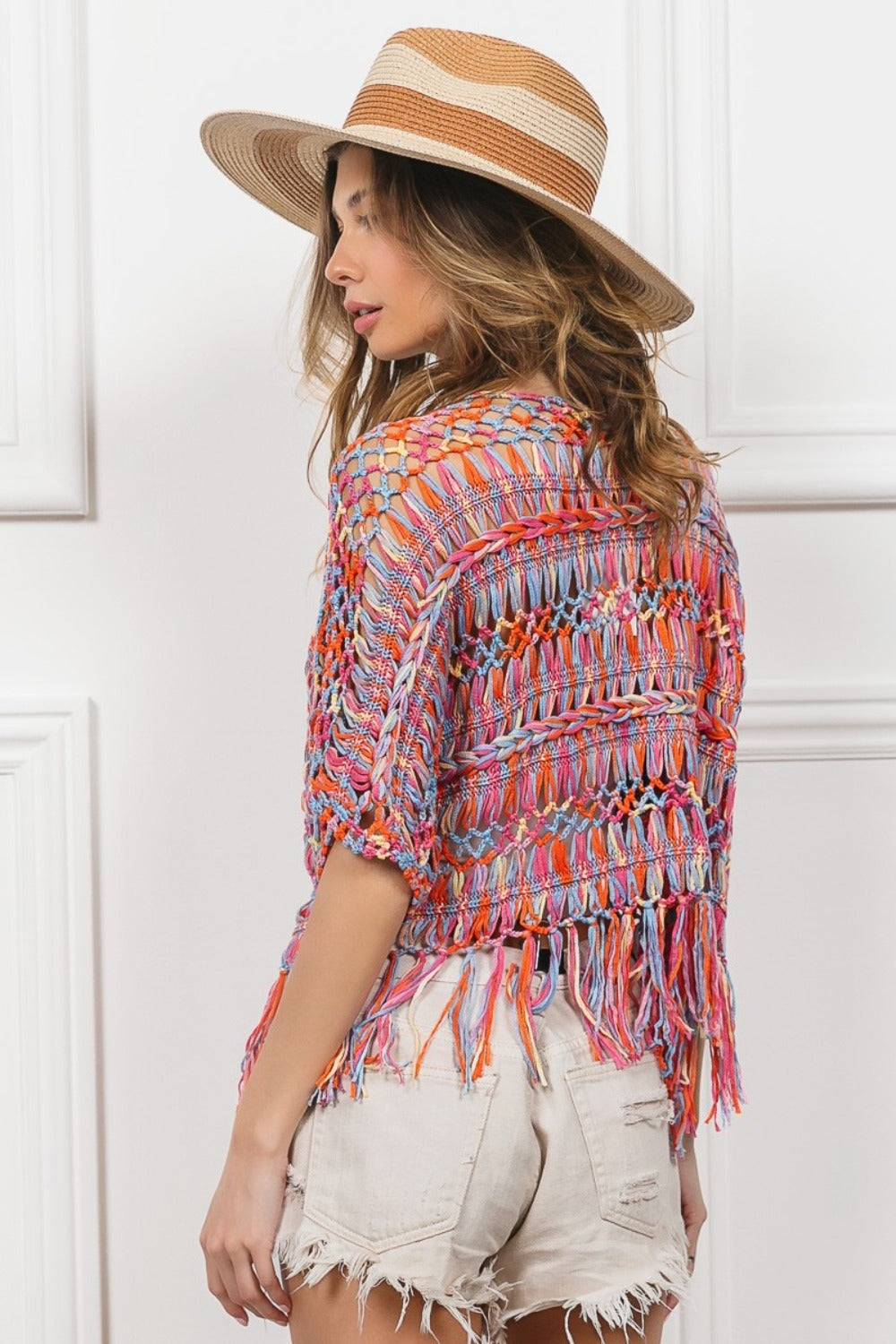 A woman, facing away against a white-paneled background, wears a BiBi Open Front Fringed Crop Knit Cardigan with distressed denim shorts and a wide-brimmed straw hat, highlighting the cardigans playful fringe.