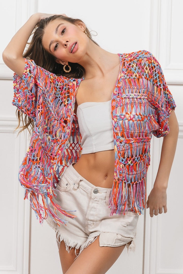 A woman confidently models the BiBi Open Front Fringed Crop Knit Cardigan in vibrant colors, layered over a white tube top and light denim shorts. Posed against a white paneled background, she exudes style with one hand on her hip and the other behind her head.