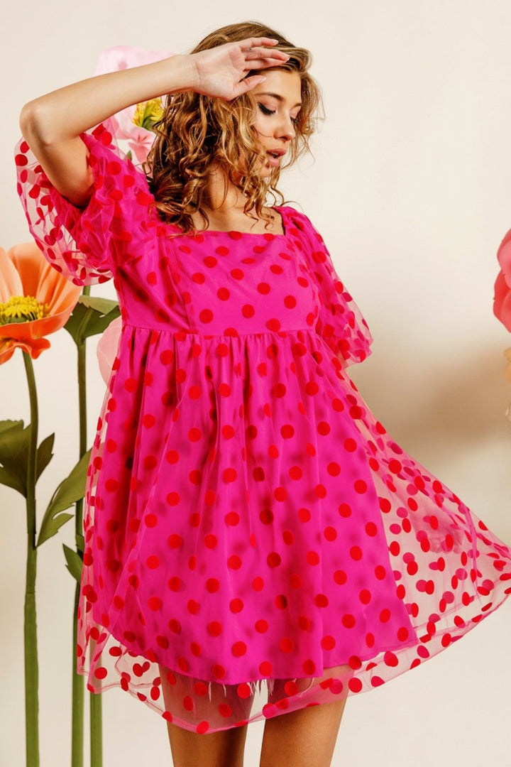 A person models the BiBi Polka Dot Mesh Puff Sleeve Dress, showcasing its bright pink hue with red polka dots, while posing gracefully in front of large, colorful flowers. The dress features vintage-inspired sheer sleeves and a flowing skirt. The softly lit background enhances the vibrant colors.