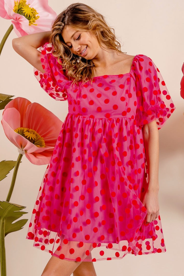 A woman models the vintage-inspired BiBi Polka Dot Mesh Puff Sleeve Dress in pink and red, striking a pose with her hand raised. This dress, featuring a square neckline, exudes charm. In the background, large artificial flowers bloom vibrantly.