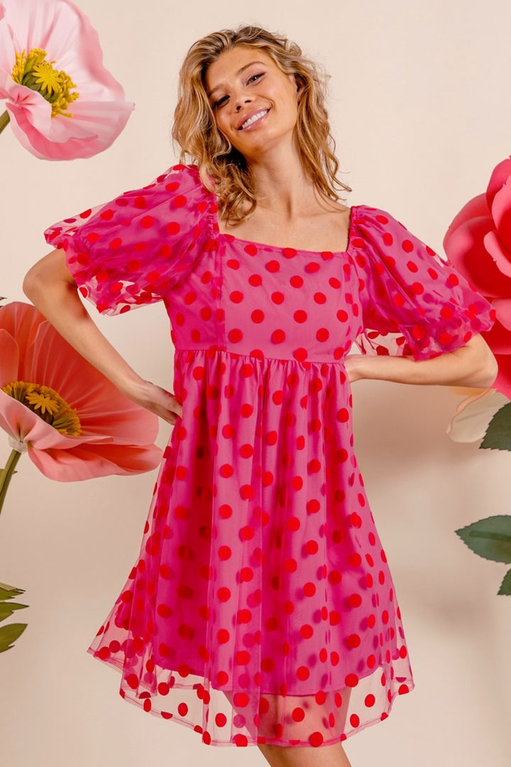 A woman in the BiBi Polka Dot Mesh Puff Sleeve Dress, vintage-inspired with a pink hue and adorned with red polka dots, stands smiling. Her dress, characterized by a square neckline, is beautifully complemented by large pink and red flower decorations set against an off-white background.