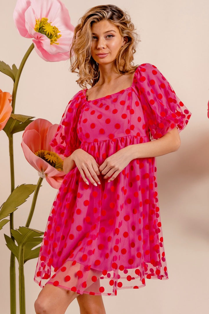 Wearing the BiBi Polka Dot Mesh Puff Sleeve Dress, a woman stands gracefully before a backdrop adorned with large, colorful flowers. Her vintage-inspired, flowing pink dress with red polka dots complements her shoulder-length wavy hair and relaxed expression as she embodies timeless elegance.