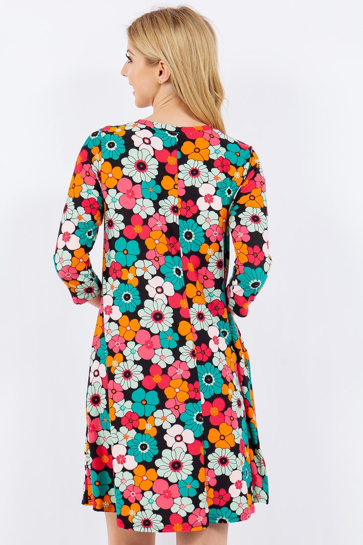 A person with long blonde hair is wearing the Celeste Full Size Floral Three-Quarter Sleeve Dress with Pockets. This vibrant summer dress showcases unique, eye-catching floral prints in orange, pink, blue, and white while the wearer faces away.