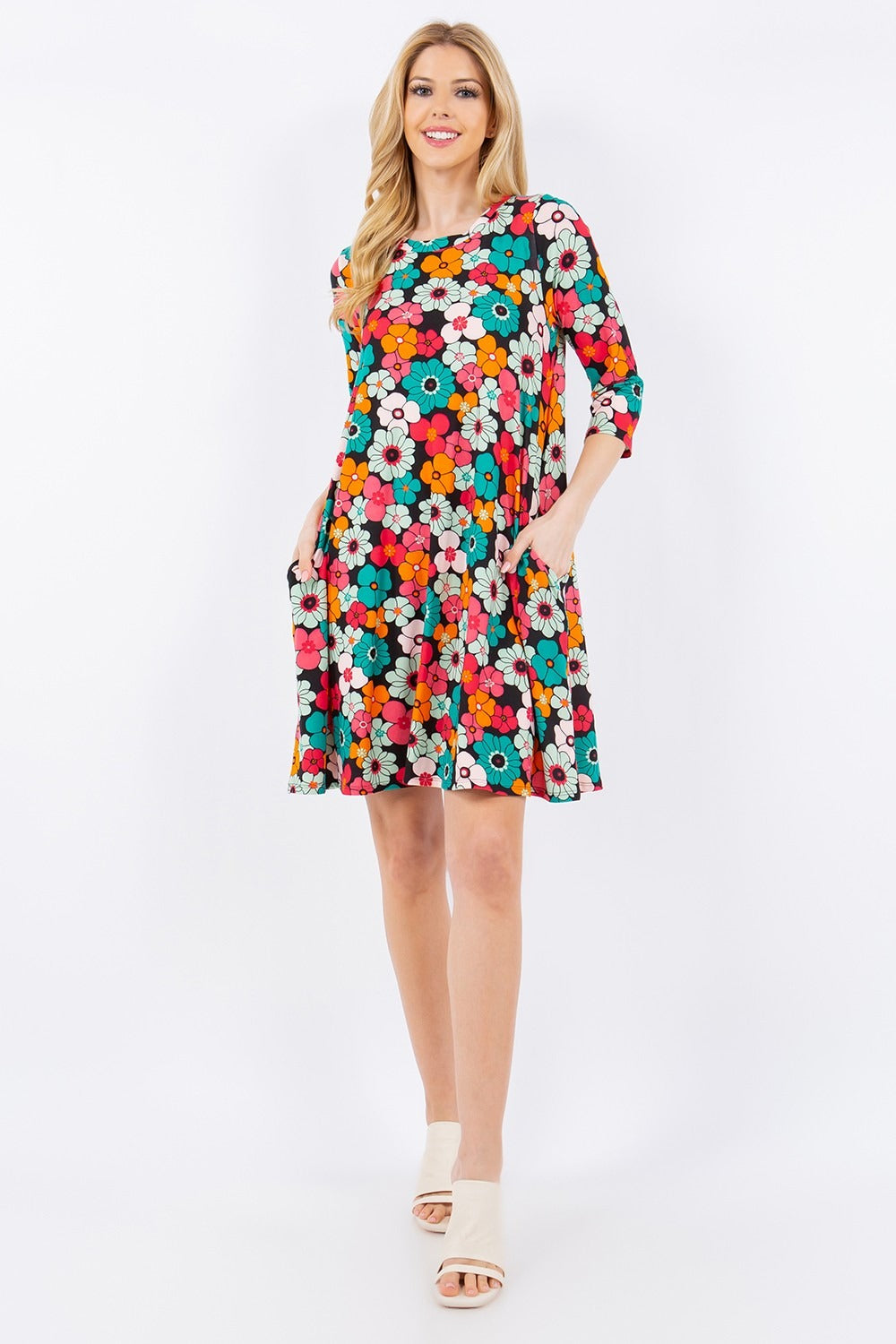 The Celeste Full Size Floral Three-Quarter Sleeve Dress with Pockets, featuring unique prints, is worn by a smiling person against a white background. They pair it with light-colored open-toed shoes.