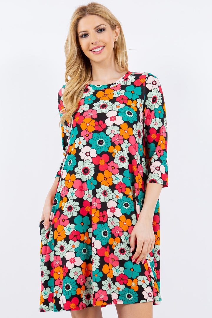 A person with long, blonde hair is wearing the Celeste Full Size Floral Three-Quarter Sleeve Dress with Pockets. The bold red, white, and teal floral pattern creates an eye-catching look as they smile against a plain white background.