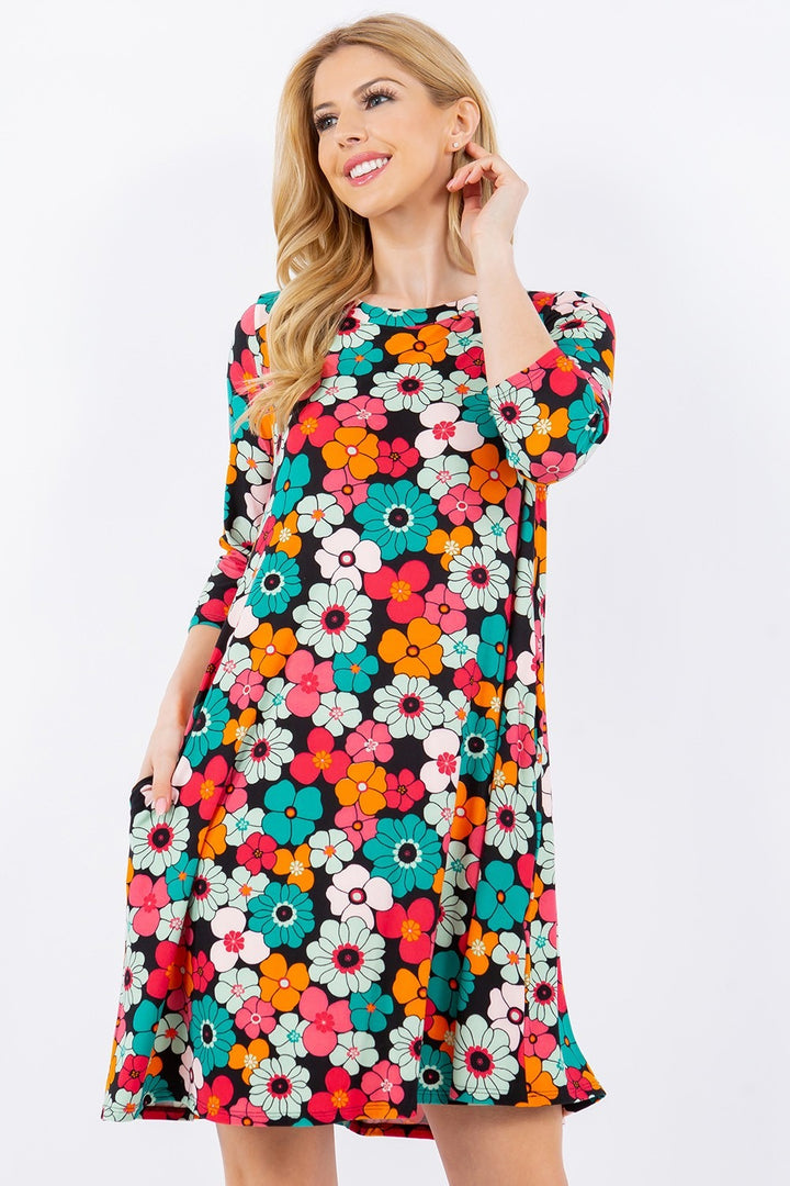 A woman smiles in the Celeste Full Size Floral Three-Quarter Sleeve Dress with Pockets, showcasing vibrant floral prints in red, orange, blue, and white. She stands against a plain background with one hand near her chin and the other in her pocket.