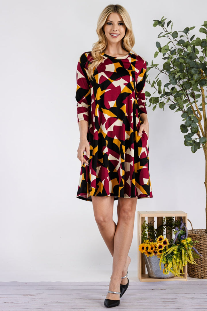 A woman smiles beside a sunflower crate and tall green plant, wearing the versatile Celeste Full Size Geometric Round Neck Dress with Pockets, featuring vibrant red, yellow, black, and white patterns. She pairs it stylishly with black shoes.