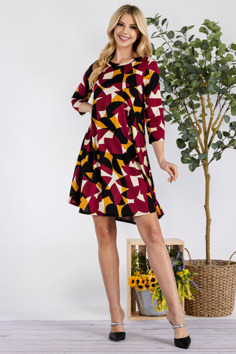 A smiling woman in a Celeste Full Size Geometric Round Neck Dress with pockets poses with one hand on her hip beside a small tree and a wicker basket filled with sunflowers.