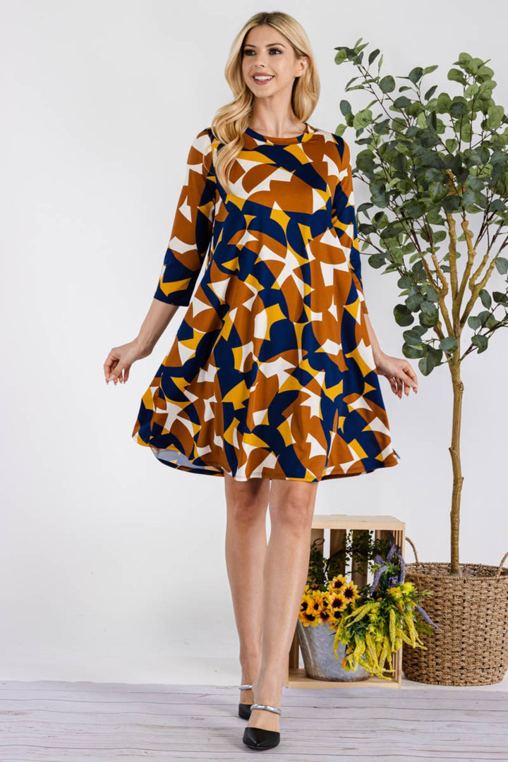 A woman joyfully poses in a Celeste Full Size Geometric Round Neck Dress with pockets, showcasing its blue, orange, and white design. Standing near a tall plant and a floor flower arrangement, she lifts the skirt playfully while smiling in black heels.