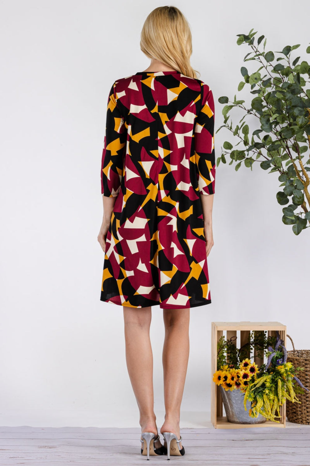 A person wearing a Celeste Full Size Geometric Round Neck Dress with Pockets featuring black, red, white, and yellow shapes stands with their back to the camera. Nearby, theres a box of sunflowers and a plant on the light wooden floor.