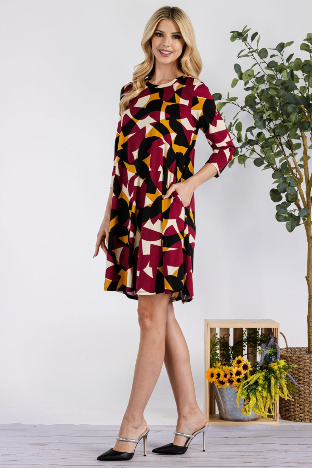 A woman in a Celeste Full Size Geometric Round Neck Dress with pockets stands smiling next to a wooden crate of sunflowers, wearing black heeled mules. Her long blonde hair cascades down as a leafy plant adorns the background.