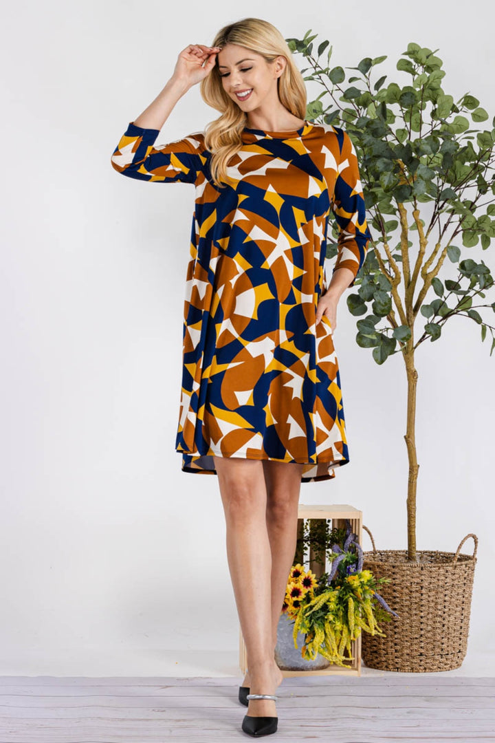 A woman in a Celeste Full Size Geometric Round Neck Dress with Pockets smiles while touching her hair, wearing black heels. She stands beside a basket containing a green plant and sunflowers, set against a plain background that emphasizes her stylish outfit.