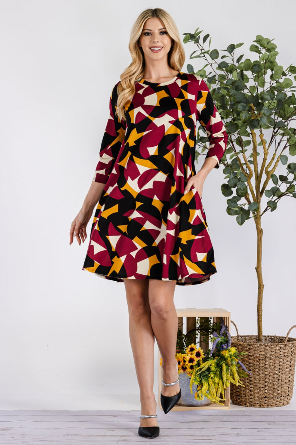 A woman in a Celeste Full Size Geometric Round Neck Dress with Pockets stands confidently by a potted plant, smiling in black heels. The multicolored design and vibrant pattern of her outfit are beautifully highlighted against the simple background.