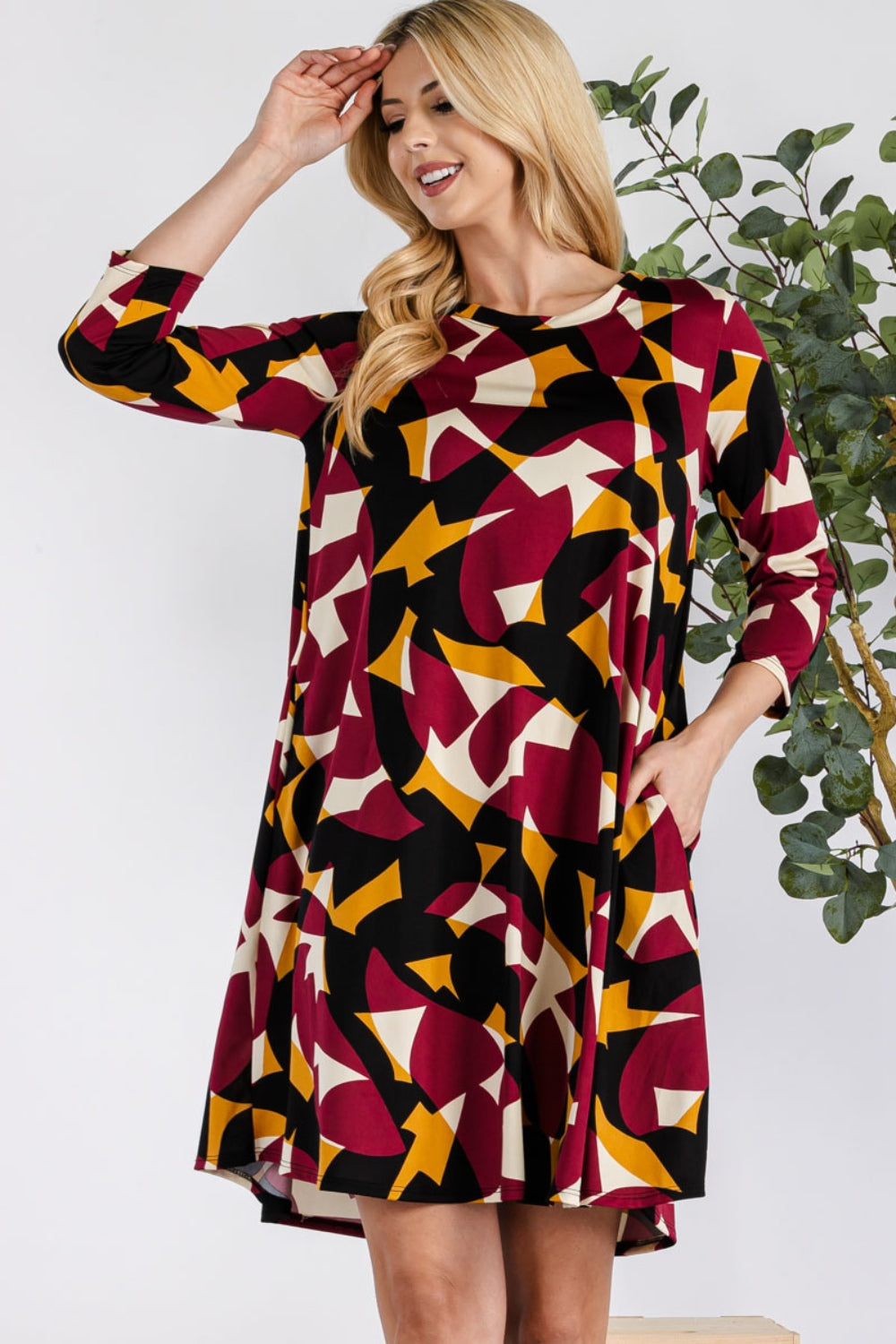 A woman smiles with one hand on her head, wearing the Celeste Full Size Geometric Round Neck Dress with Pockets, featuring vibrant red, yellow, and black shapes. This versatile dress fits any occasion, with a lush green plant as the backdrop.