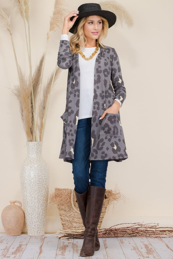 A person wearing a black hat, the Celeste Full Size Leopard Open Front Contrast Cardigan, a white shirt, jeans, and tall brown boots stands next to a large vase with pampas grass. The light beige background features decorative baskets on the floor.