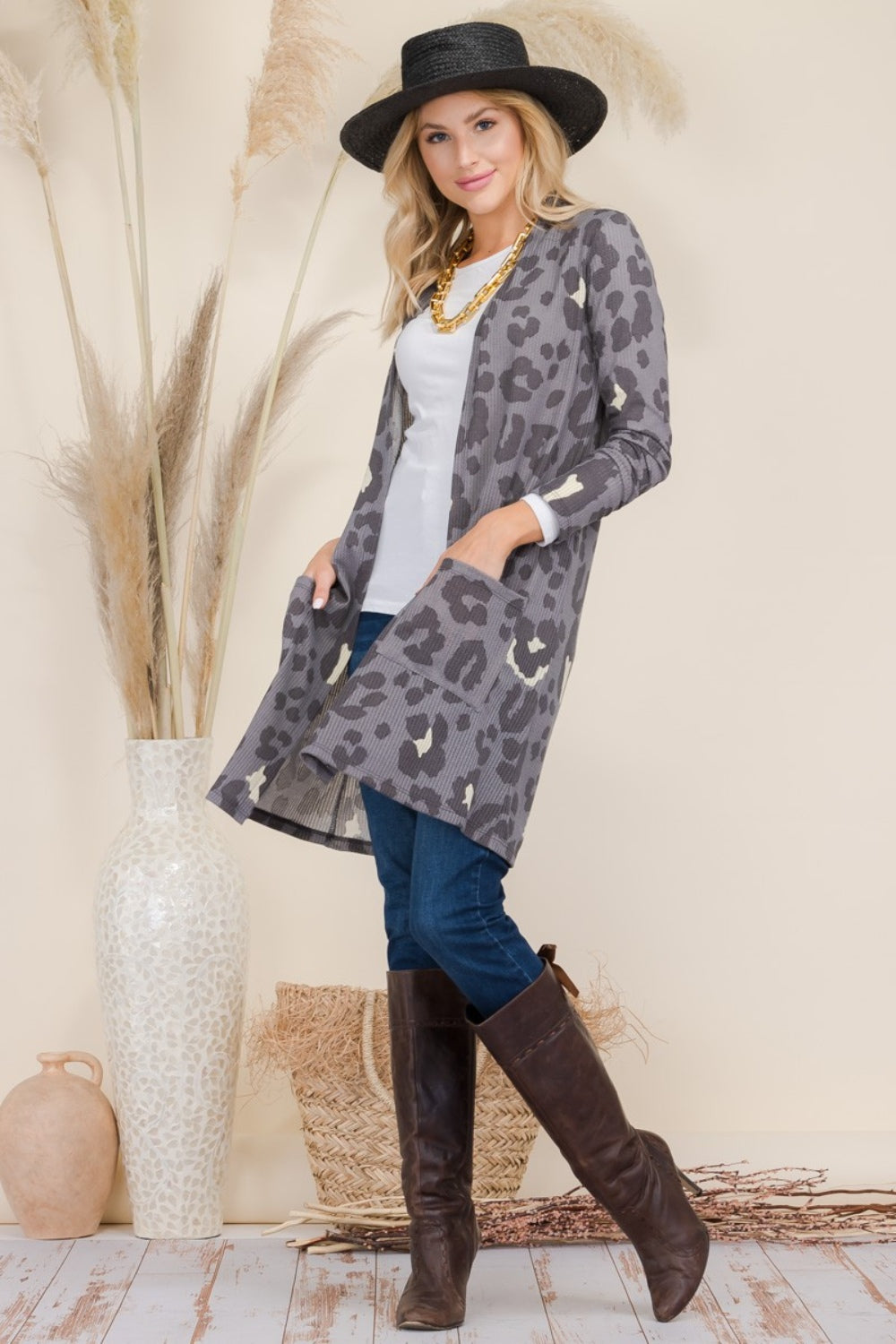 A woman effortlessly poses indoors wearing a Celeste Full Size Leopard Open Front Contrast Cardigan, a white shirt, blue jeans, a wide-brimmed black hat, and brown knee-high boots beside a large vase with decorative grass.