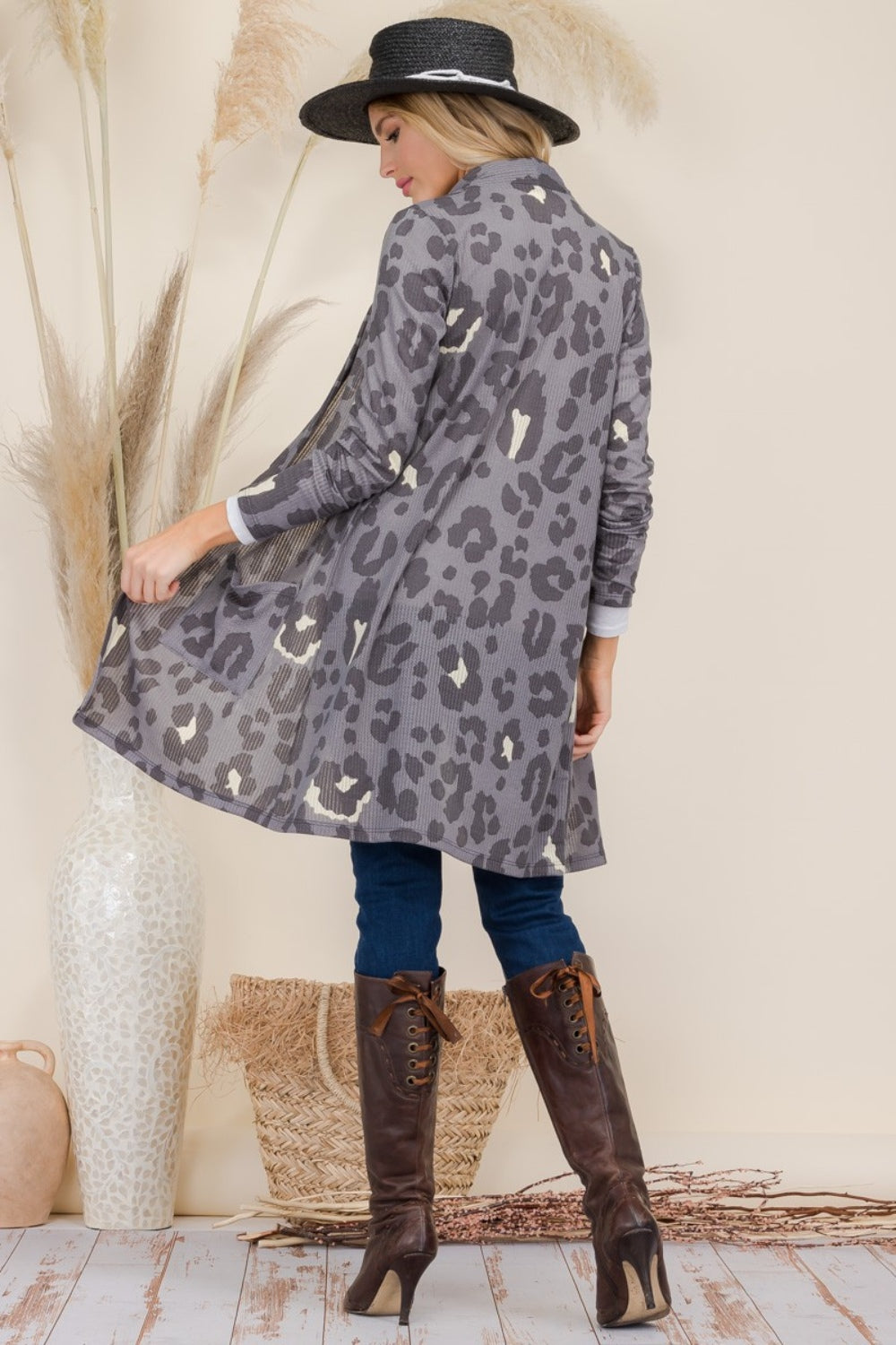 A person in a black hat and the Celeste Full Size Leopard Open Front Contrast Cardigan poses with their back to the camera. Theyre wearing blue jeans and tall brown boots, with dried plants in vases in the background.