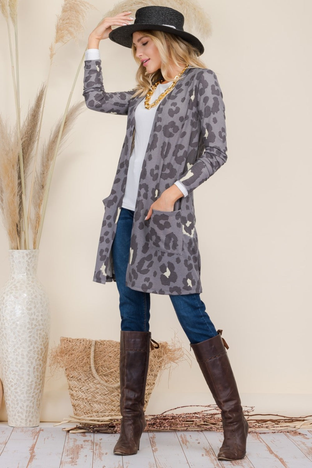 A woman in a Celeste Full Size Leopard Open Front Contrast Cardigan, white top, blue jeans, and brown knee-high boots stands in profile. She adjusts her black hat beside a large vase with pampas grass against a beige background.